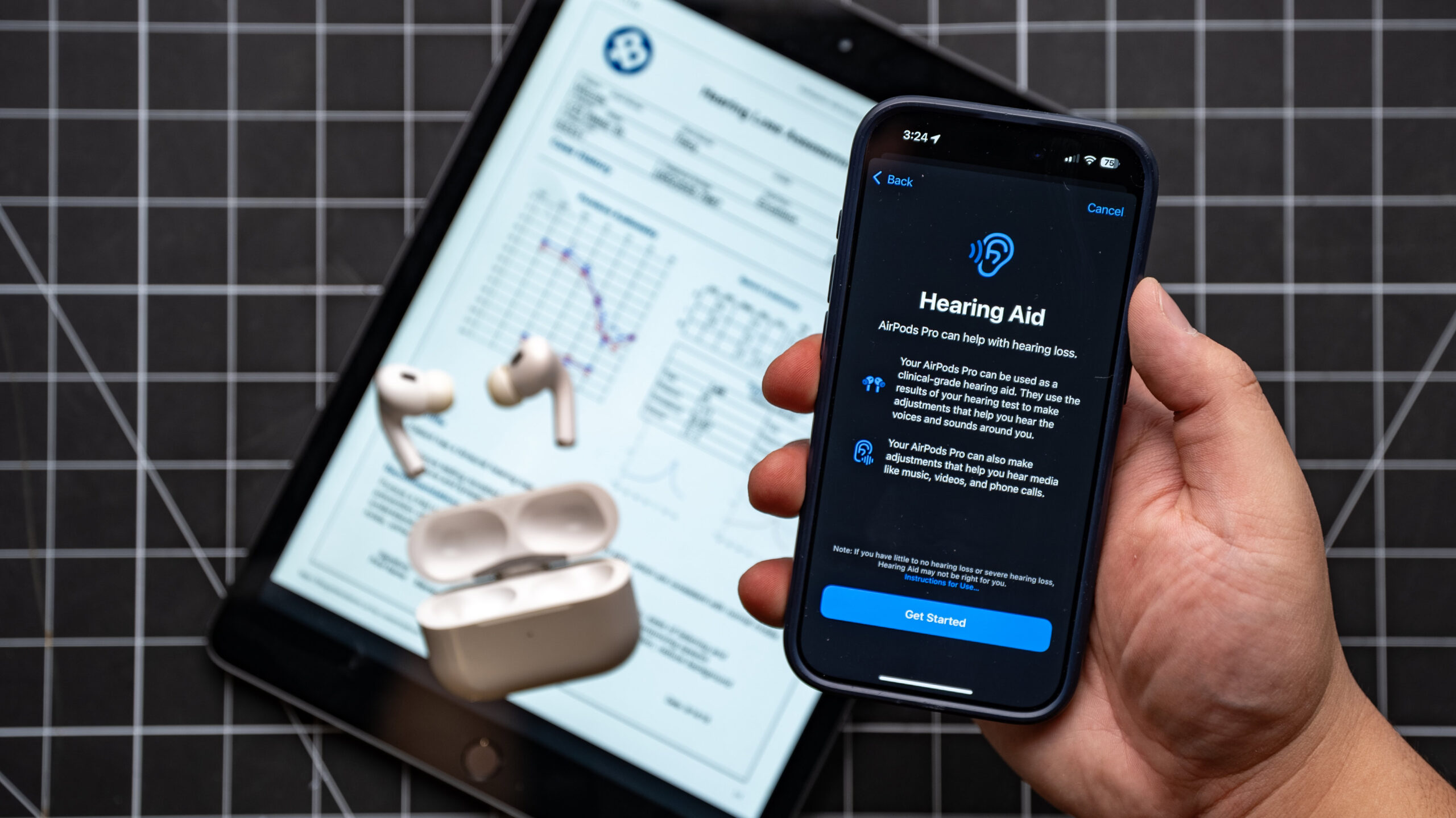 airpods pro 2 hearing aid with test results in background iPhone in hand showing Hearing Aid setup screen with AirPods Pro 2 on table on top of iPad showing audiogram hearing test results