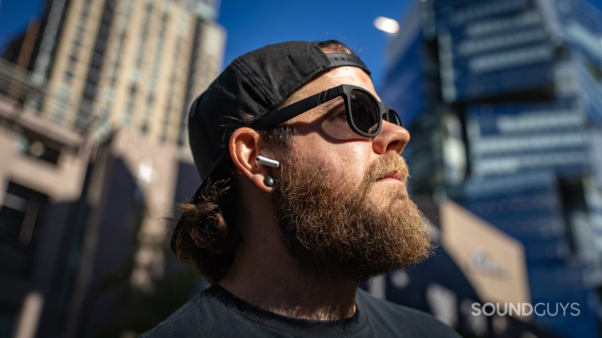 Apple AirPods 4 side profile man wearing outside library 4 A man wearing an AirPods 4 earbud outside in the sunlight surrounded by buildings.
