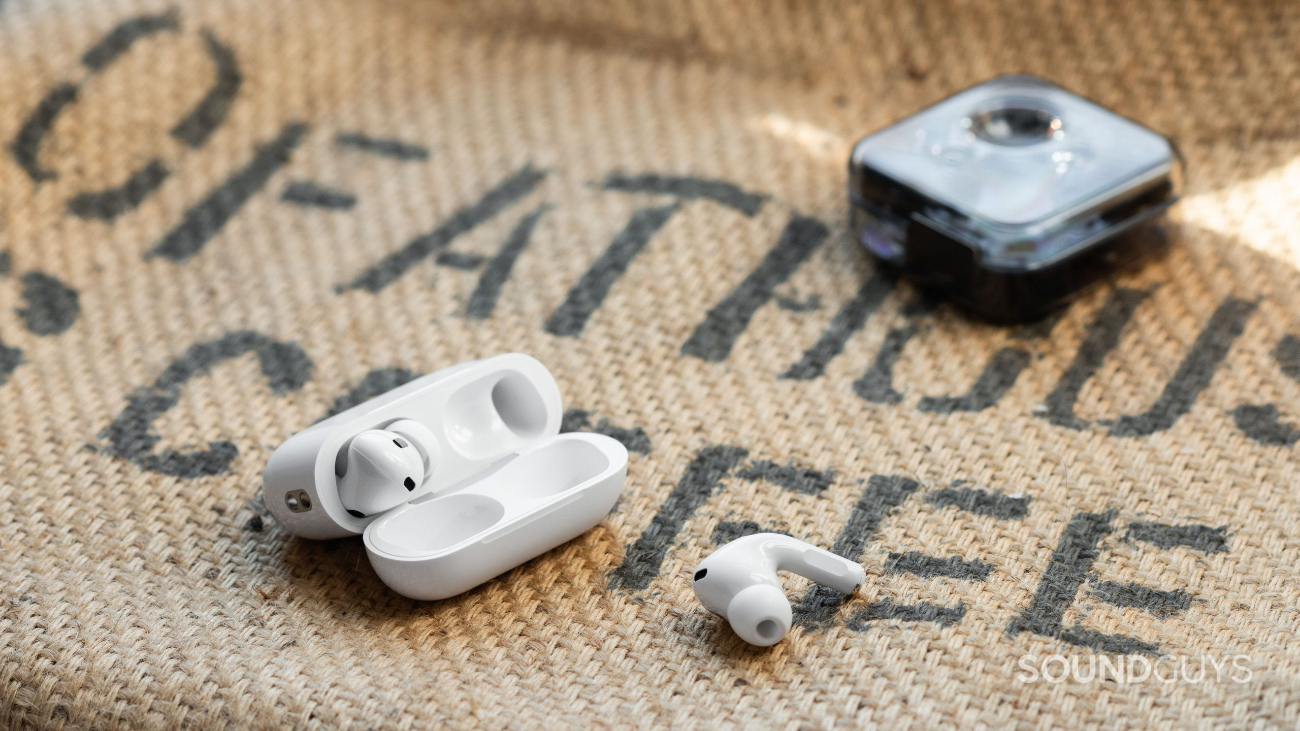Apple AirPods Pro (2nd generation) true wireless earbuds nothing ear 1 The Apple AirPods Pro (2nd generation) case open with one of the earbuds on a burlap sack, and the Nothing Ear 1 case in the background.