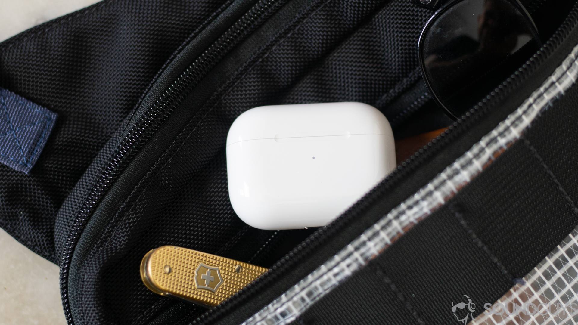 Apple AirPods Pro-5 The Apple AirPods Pro charging case next to a gold swiss army knife in the pocket of a bag.