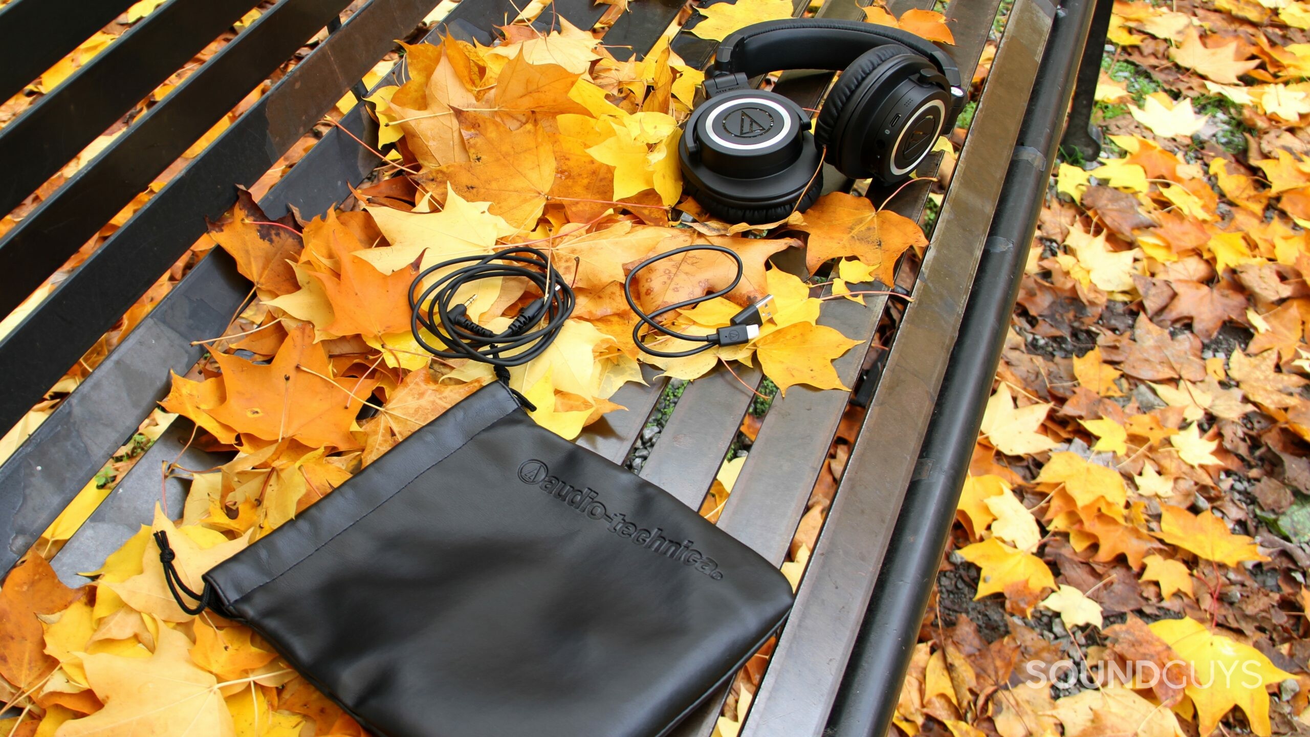 audio-technica-ath-m50xbt2-accesories Audio-Technica ATH-M50xBT2 with pouch, USB cable, and headphone jack cable on a bench.
