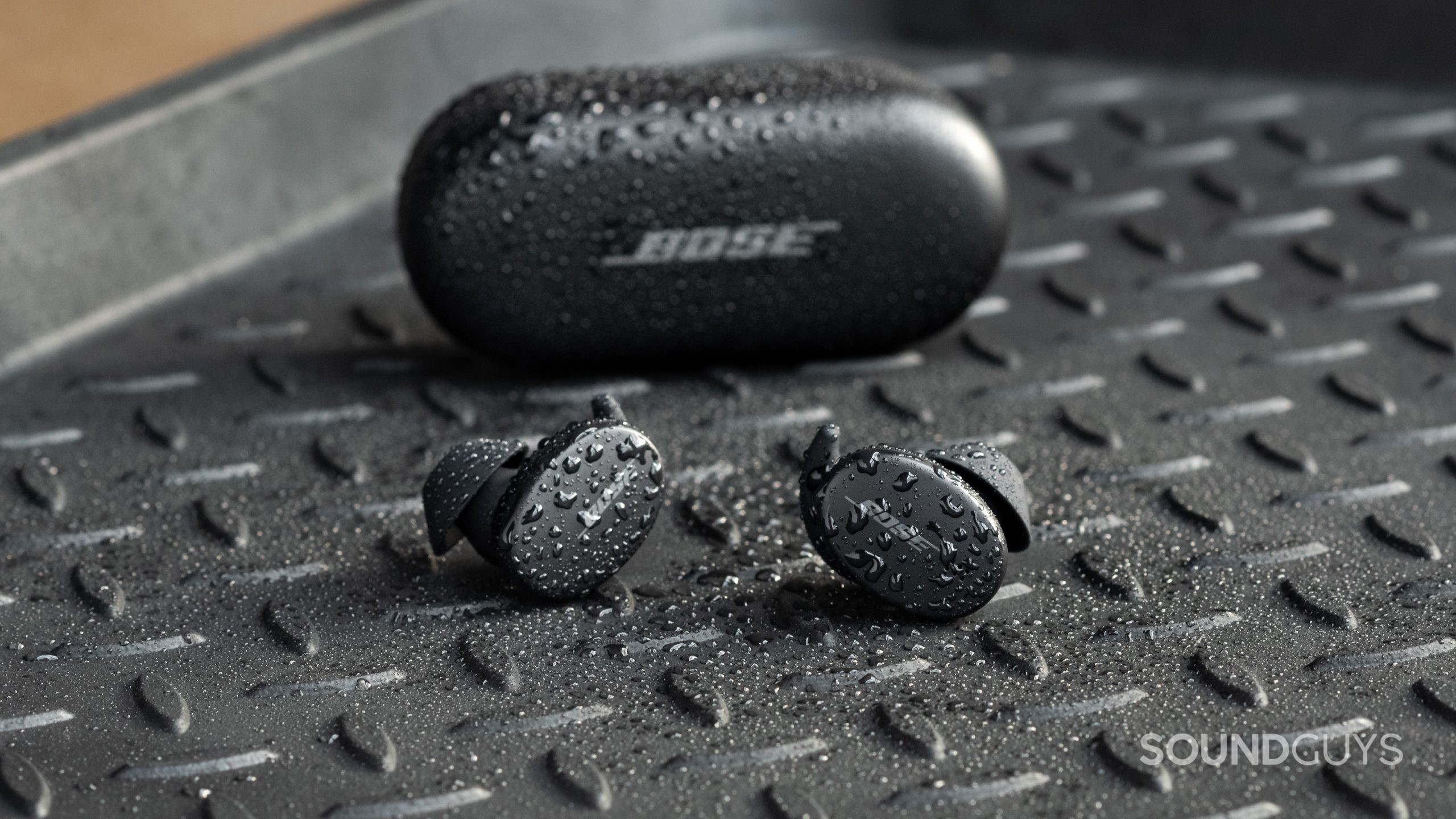 Bose Sport earbuds water The Bose Sport Earbuds true wireless workout earbuds sit outside of the closed charging case, all objects are covered in sprinkles of water.