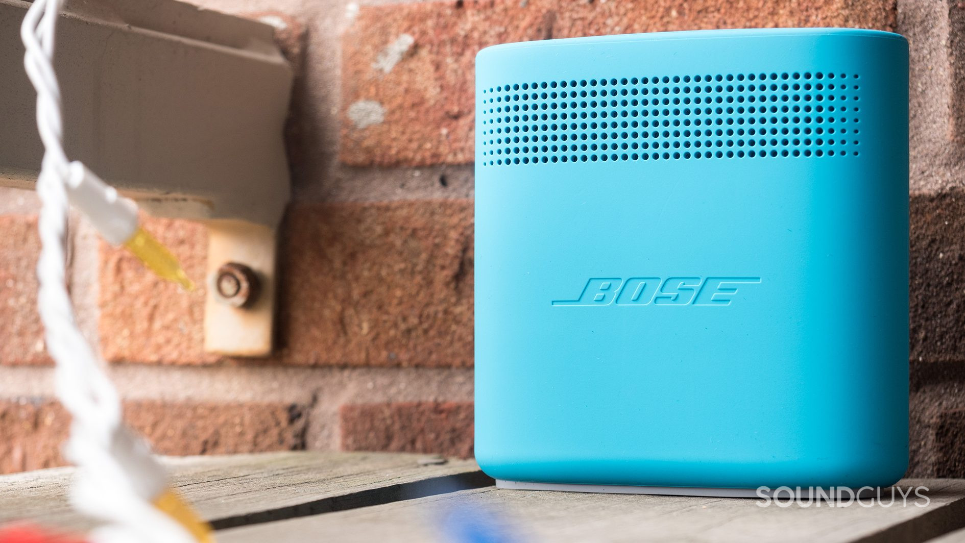 Bose_SoundLink Color II_LK_5 Dual-opposing, passive radiators are housed inside the Bose SoundLink Color II speaker. This minimizes surface vibrations while maximizing volume output. Pictured: The back of the Bose SoundLink Color II speaker, which has dotted openings on top-forth of the panel and the Bose logo emblazoned in the middle. It is ona wood balcony with decorative lights and a brick backdrop.