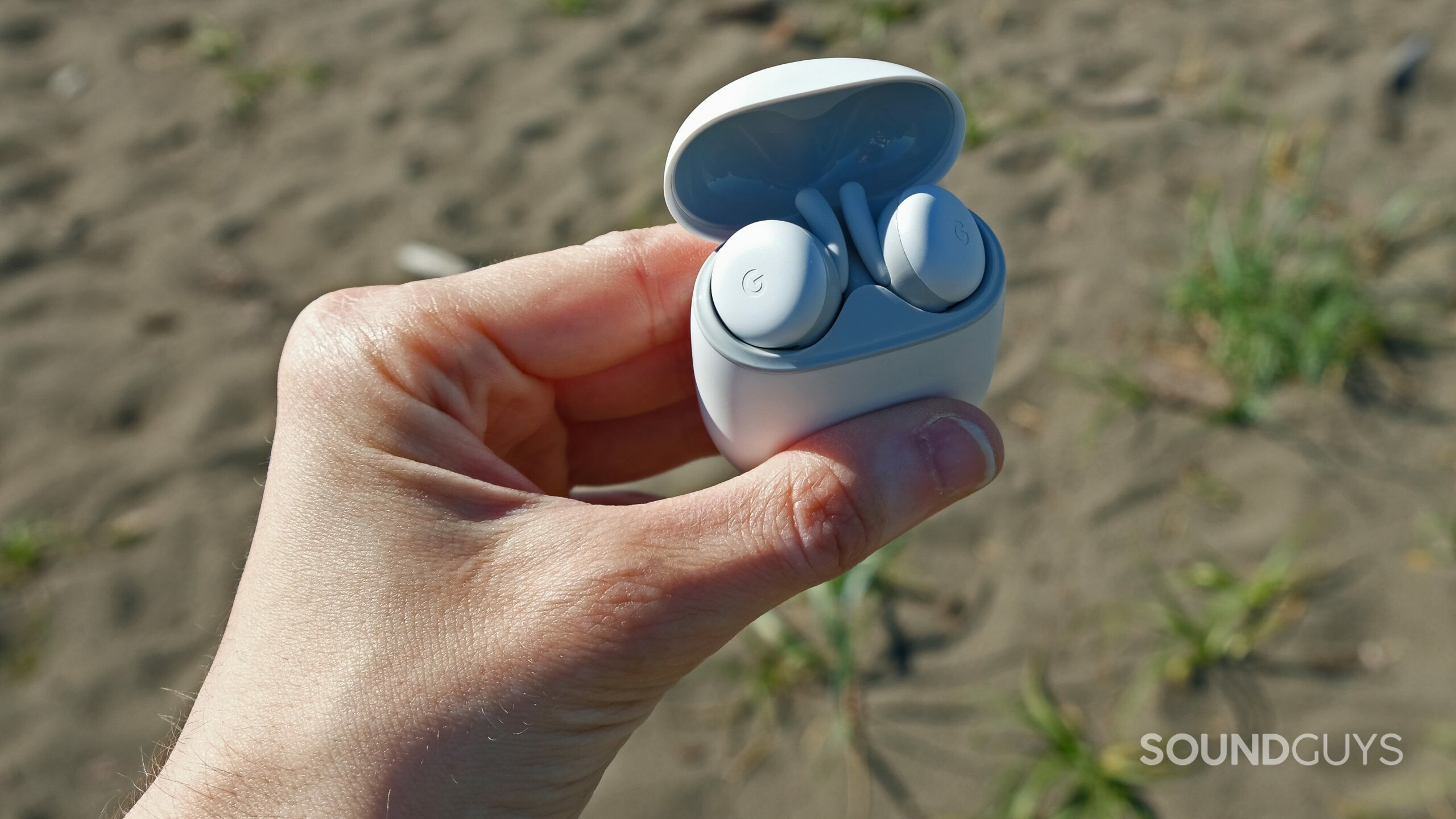 google pixel buds a-series hand holds A hand holds the open case of the Google Pixel Buds A-Series in front of a beach.