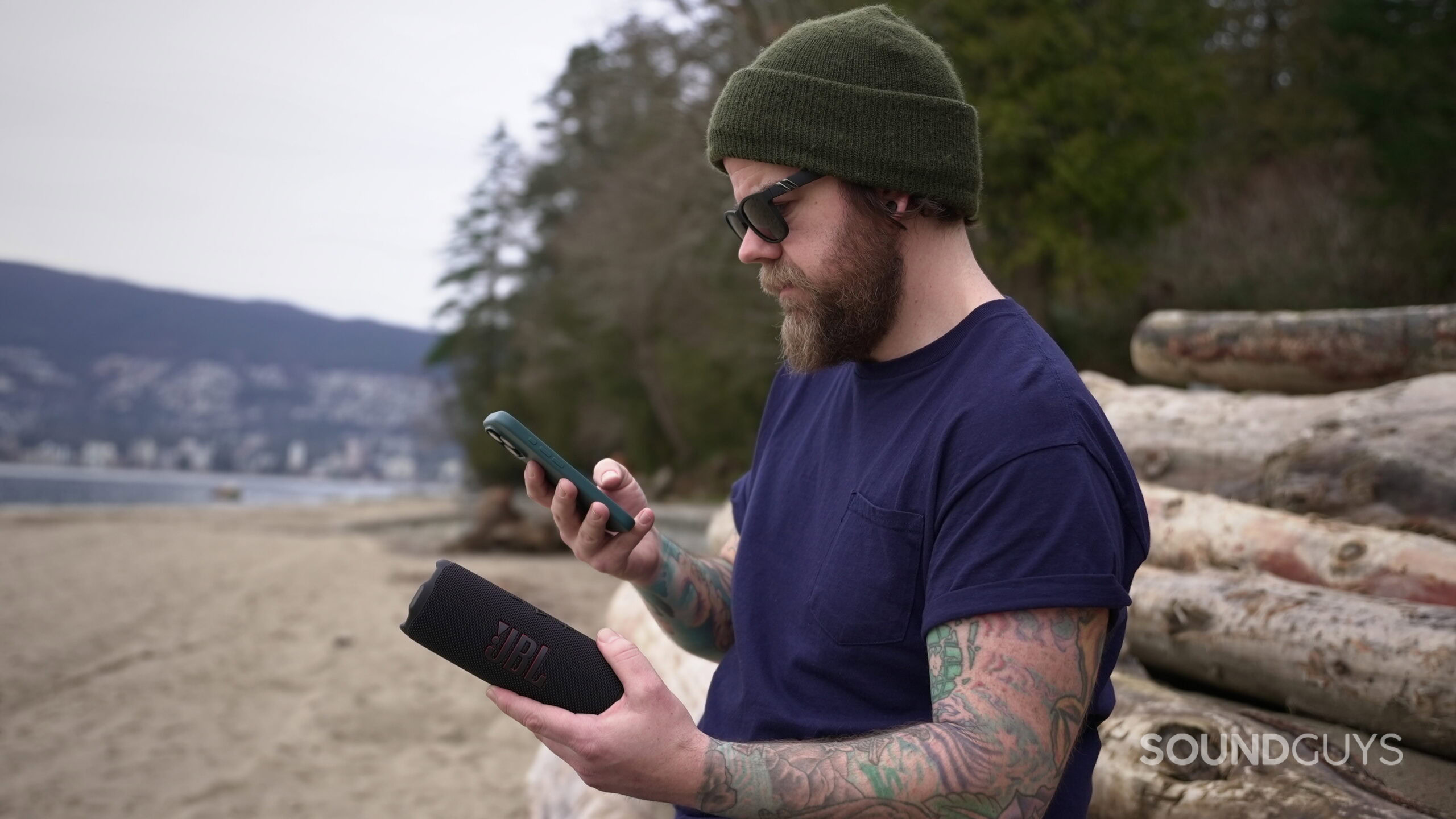 Flip 7 at the beach - 1 A man at the beach holding a Flip 7 speaker in his left hand, and his phone in his right hand.