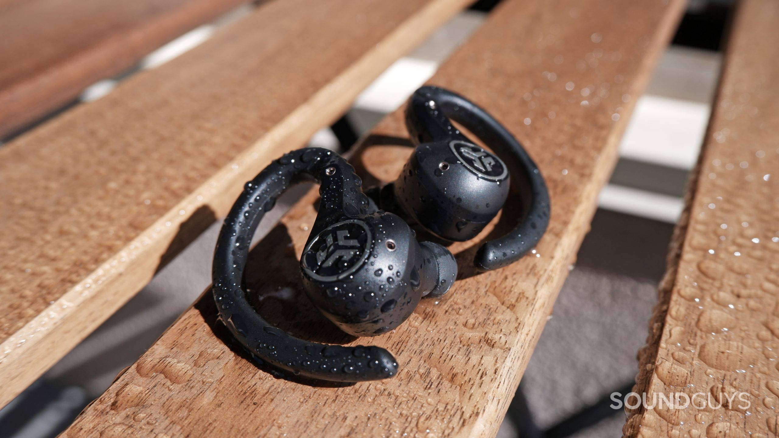 jlab-epic-air-sport-anc-waterproof The JLab Epic Air Sport ANC on a wooden table ouside, with water droplets on the earbuds and table.