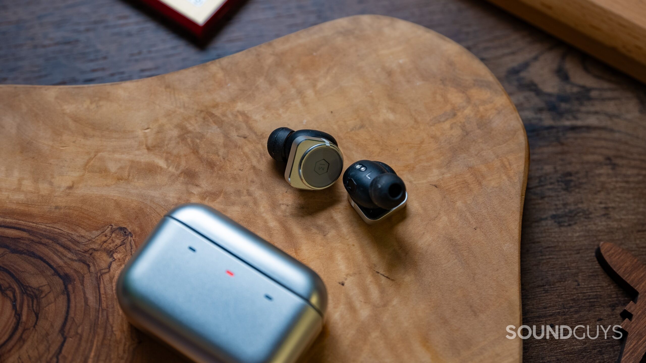Master and Dynamic MW09 Hero Image #4 Master & Dynamic MW09 earbuds next to their charging case with an LED light on the charging case with the color red.