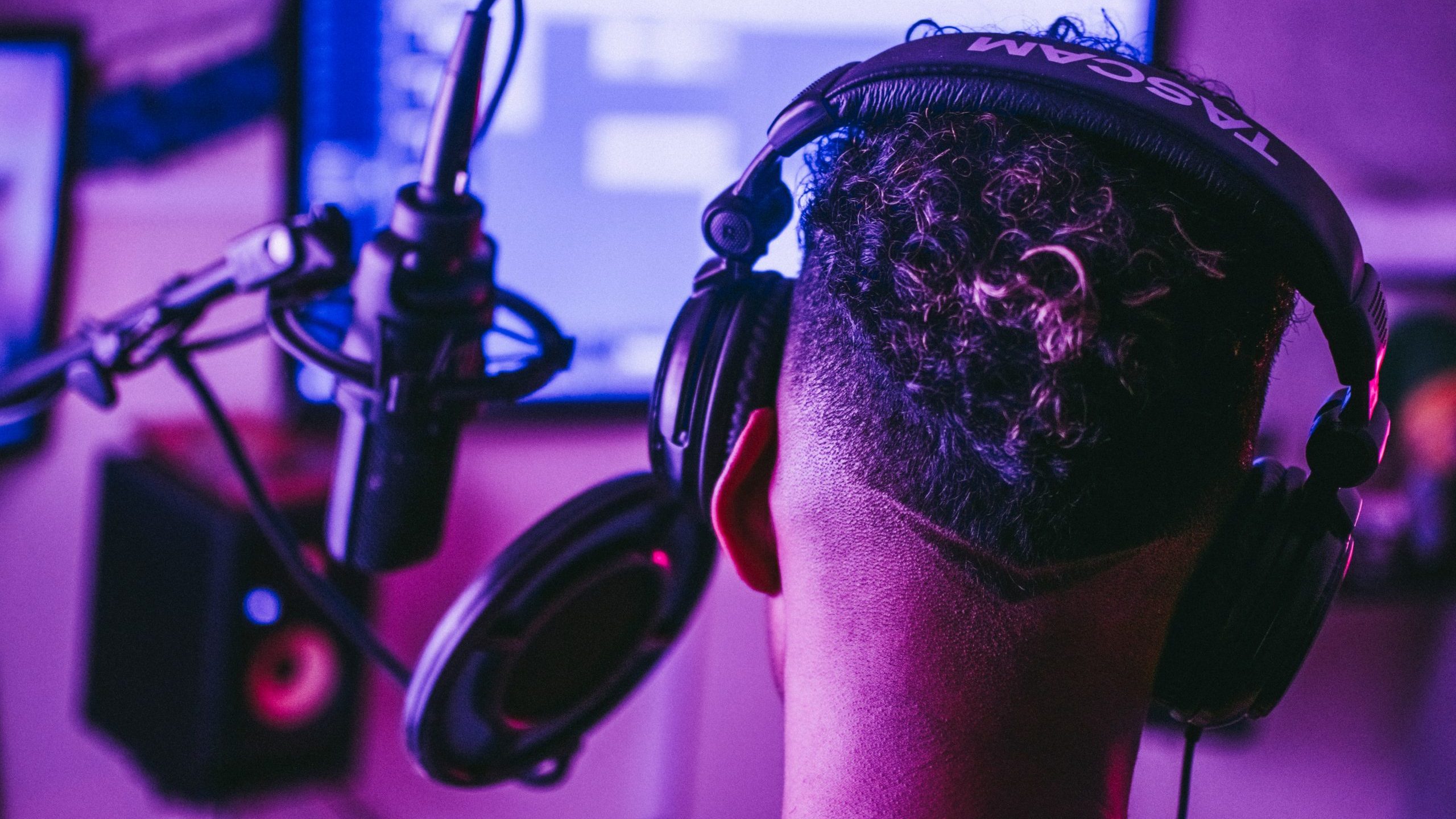 pexels-davis-sanchez-3916376 The back of a person's head wearing headphones positioned in front of a recording microphone. In the background the interface of a DAW is visible.