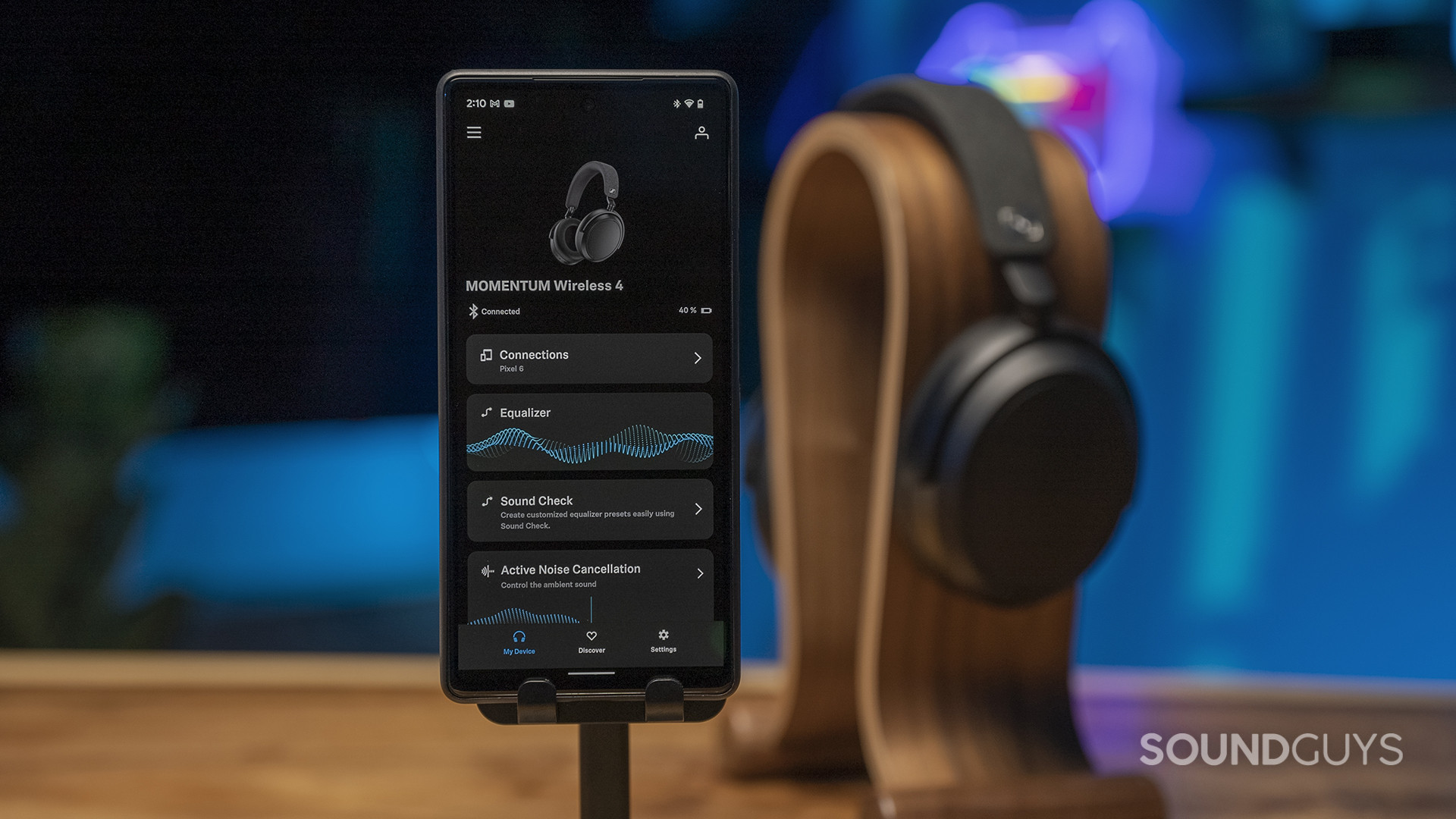sennheiser-momentum-4-wireless-app The Sennheiser Momentum 4 Wireless sits on a headphone stand in background with a phone displaying the Sennheiser Smart Control app displayed.